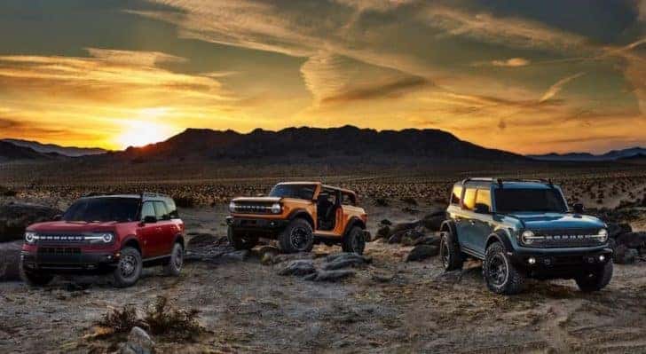 A red 2021 Ford Bronco Sport, a yellow Bronco 2-door with no roof or doors and a blue Bronco 4-door are parked in a rocky desert at sunset, soon to be seen at a Ford Bronco dealer in Canon City.