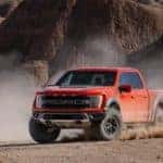 An orange Ford F-150 Raptor kicks up dust in the desert against a backdrop of sand dunes