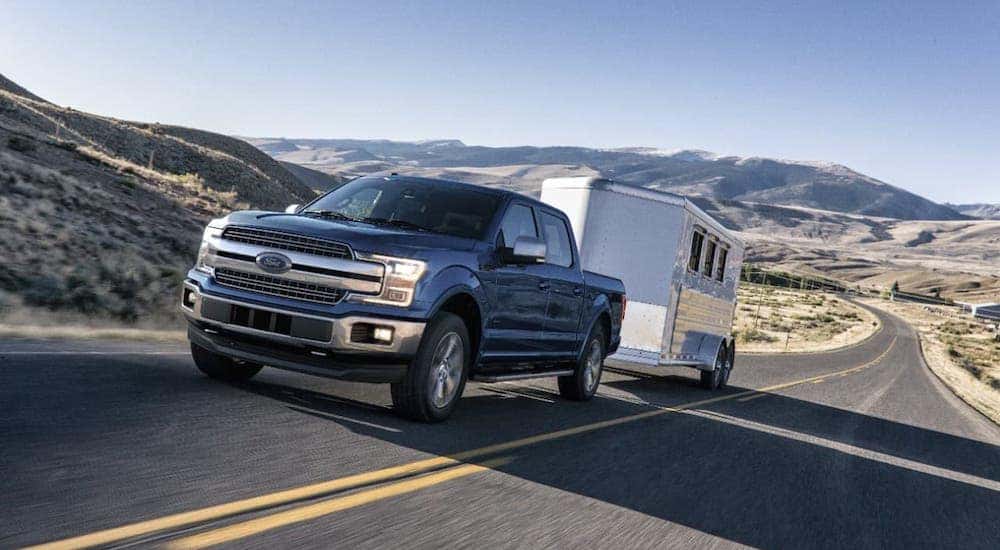A blue Ford F-150 pickup truck tows a trailer as it climbs a mountain highway