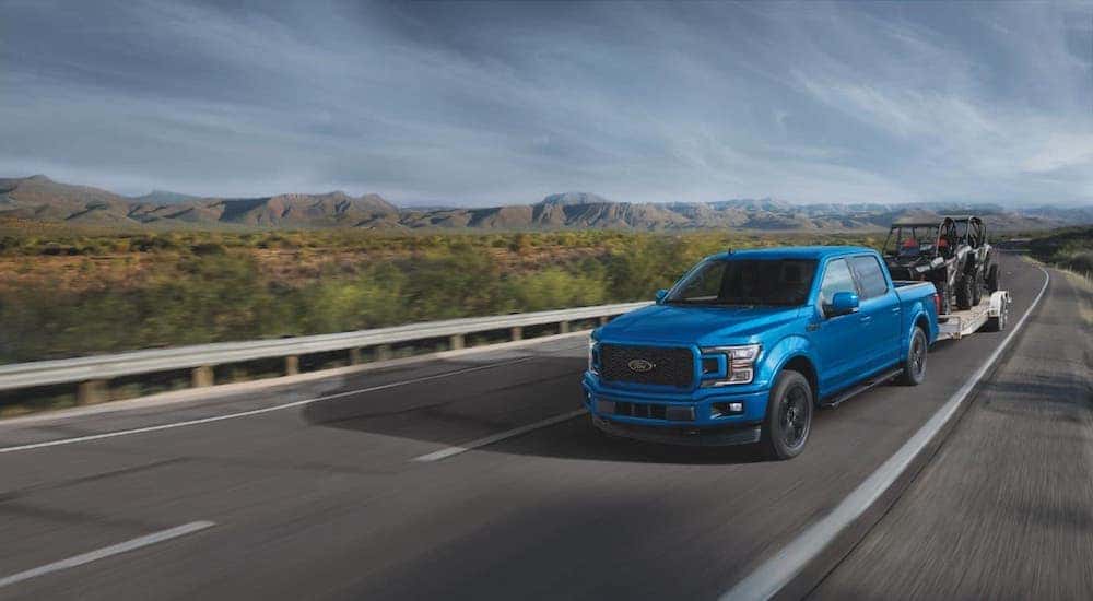 A blue 2019 Ford F-150 towing a trailer with ATVs on an empty highway.