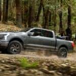 Side view of a silver 2022 Ford F-150 Lightning driving through the woods after leaving a Canon City car dealership.