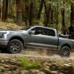 Side view of a grey 2022 Ford F-150 Lightning off-roading through the forest with a dirt bike in its bed.