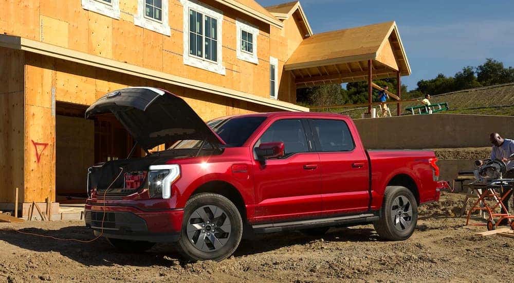 A red Ford F-150 Lightning in front of a construction site; the hood is popped, with power cables running to nearby equipment
