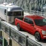 A red 2024 Ford F-150 Sport is shown towing a trailer across a snowy bridge after visiting a dealer with a Ford F-150 for sale near Pueblo West.