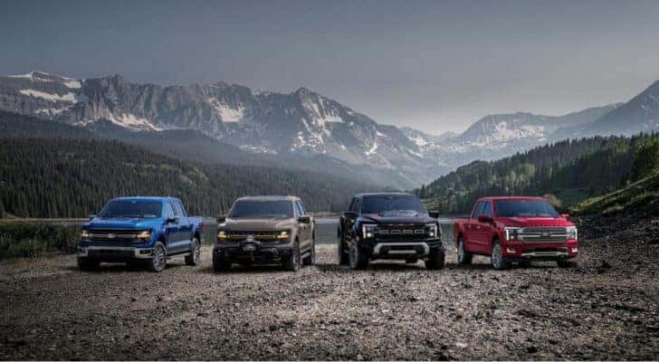 A blue 2025 Ford F-150, a brown Tremor, a black Raptor, and a red Platinum are shown overlooking mountains.