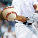 Close-up of a brown bat held by a man in white pants and a white shirt hitting a baseball