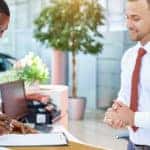 Man at the dealership front desk signing papers on a clipboard with a man in a white shirt and red tie standing by for help