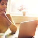 Woman in a yellow sweater sitting at her desk using a laptop with her hand on her chin