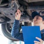 Man in a blue shirt and hat holding a pen and a blue clipboard underneath a lifted vehicle
