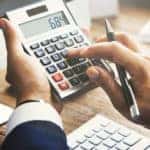 Hands of a man in a suit holding a calculator in front of a computer on a brown desk