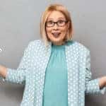 Blonde woman with glasses in a blue shirt and blue and white polka dot cardigan shrugging with cash