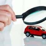 A hand holding a magnifying glass over a red toy car, reflective white background