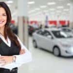 A sales woman is standing in front of cars in a showroom.