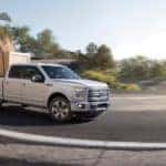 A white 2015 Ford F-150 is driving around a corner while towing a crate.