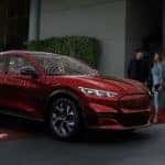 A couple is standing next to their red 2021 Ford Mustang Mach-E that's parked under accent lights at night.