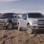 A black and a silver 2018 Ford F-150 are parked off-road in the dirt with distance mountains behind them.