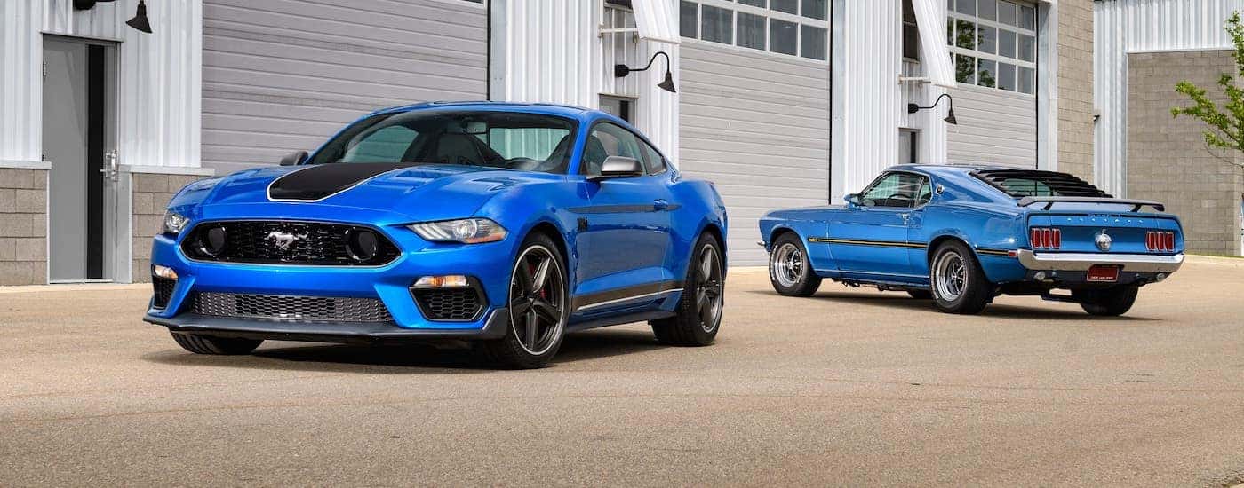 A blue 2021 Ford Mustang Mach 1 and a blue 1970s Mustang Mach 1 are parked in front of a garage.