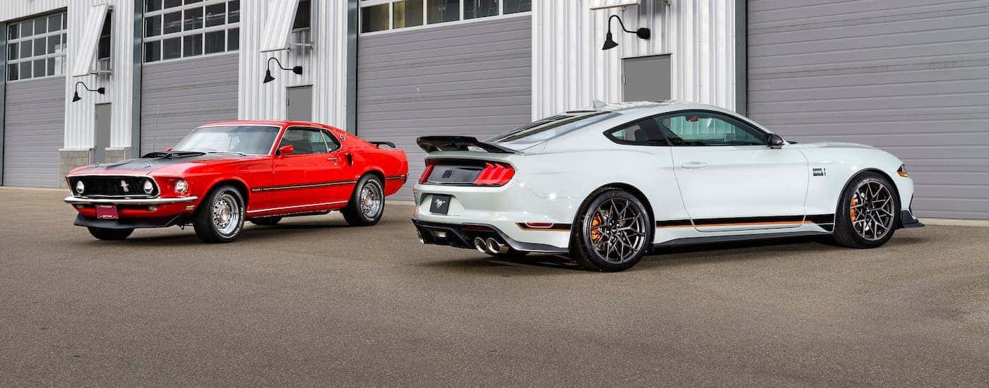 A white 2021 Ford Mustang Mach 1 and a red 1970s Mach 1 are parked in front of a garage.