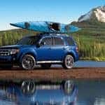 A blue 2010 Ford Escape with a kayak on the roof is parked in front of a river and snow-covered mountain.