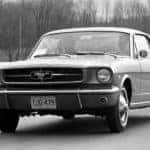 A black and white photo is showing a 1964 Ford Mustang driving down the road.