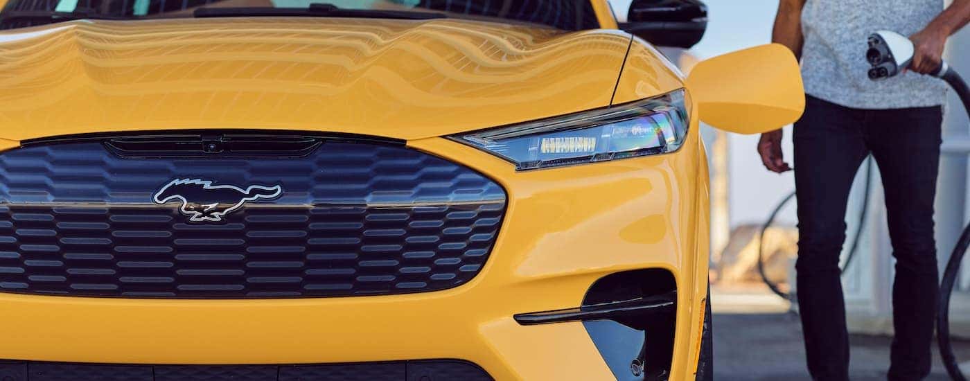 A close up shows the grille and driver side headlight of a yellow 2021 Ford Mustang Mach-E at a charging station.