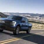 A blue 2022 Ford F-150 Lightning is shown from the front at an angle while it drives down a desert road.