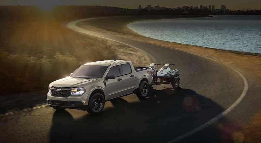 A gray 2024 Ford Maverick is shown towing two jet skis near a lake.