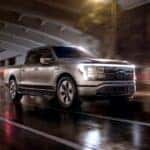 A silver 2024 Ford F-150 Lightning driving on a wet city road at night.