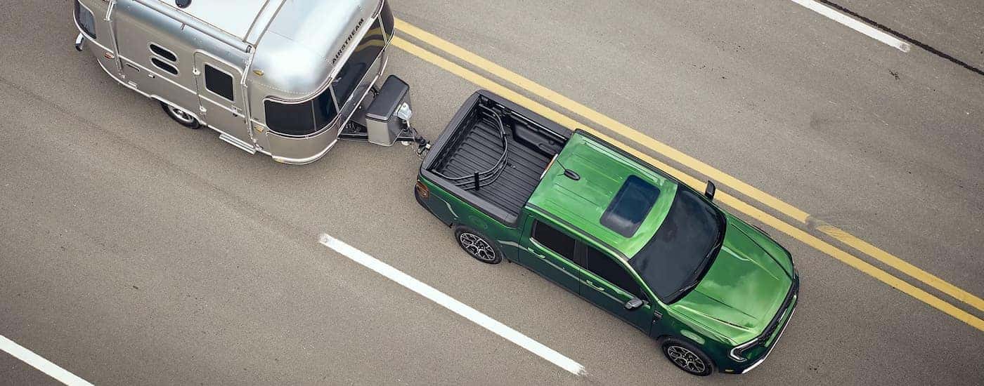 A green 2025 Ford Maverick towing a silver camper on a highway.