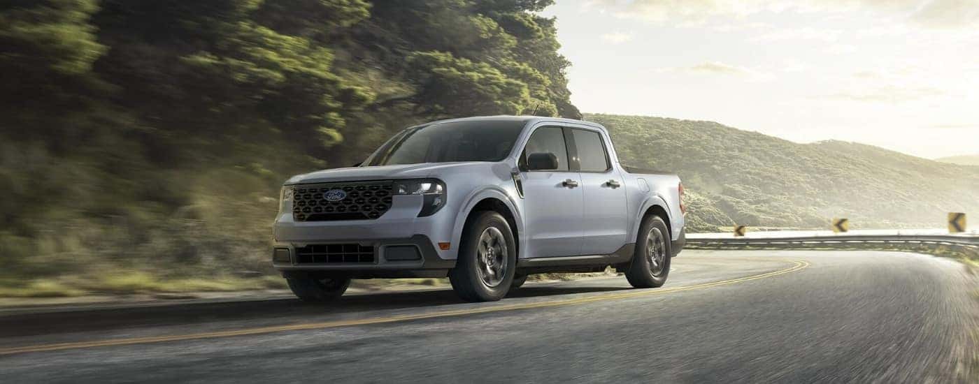 A grey 2025 Ford Maverick for sale is driving on a tree-lined road.