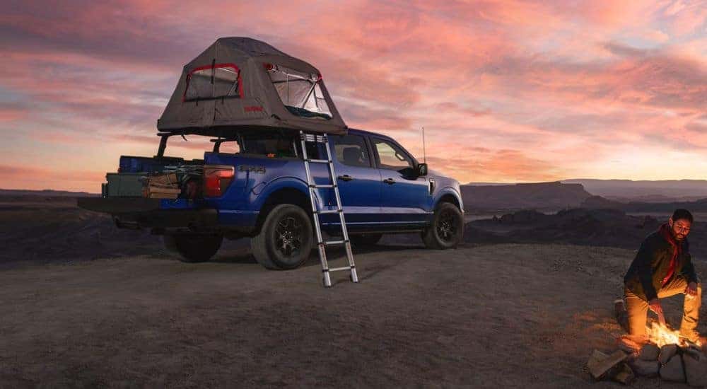 A person tending a fire next to a blue 2026 Ford F-150 STX FX4 with a tent accessory.
