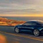 A blue 2021 Ford Mustang is shown from the side driving on a highway during a sunset.