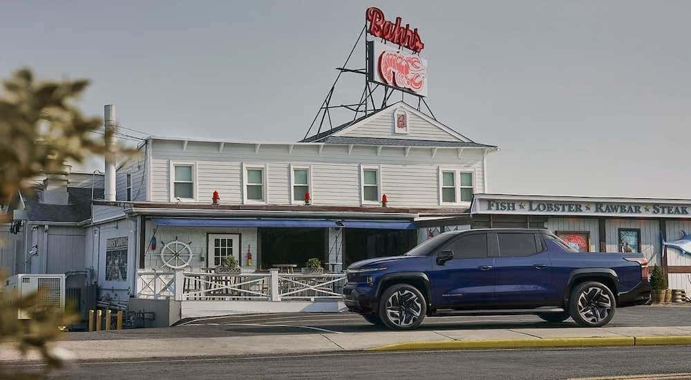 A blue 2024 Chevy Silverado EV is shown parked outside a seafood restaurant. 