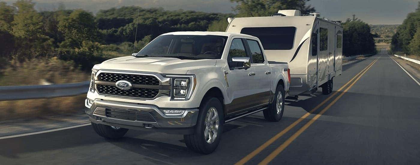 A white 2023 Ford F-150 XLT is shown towing a camper on an open road.