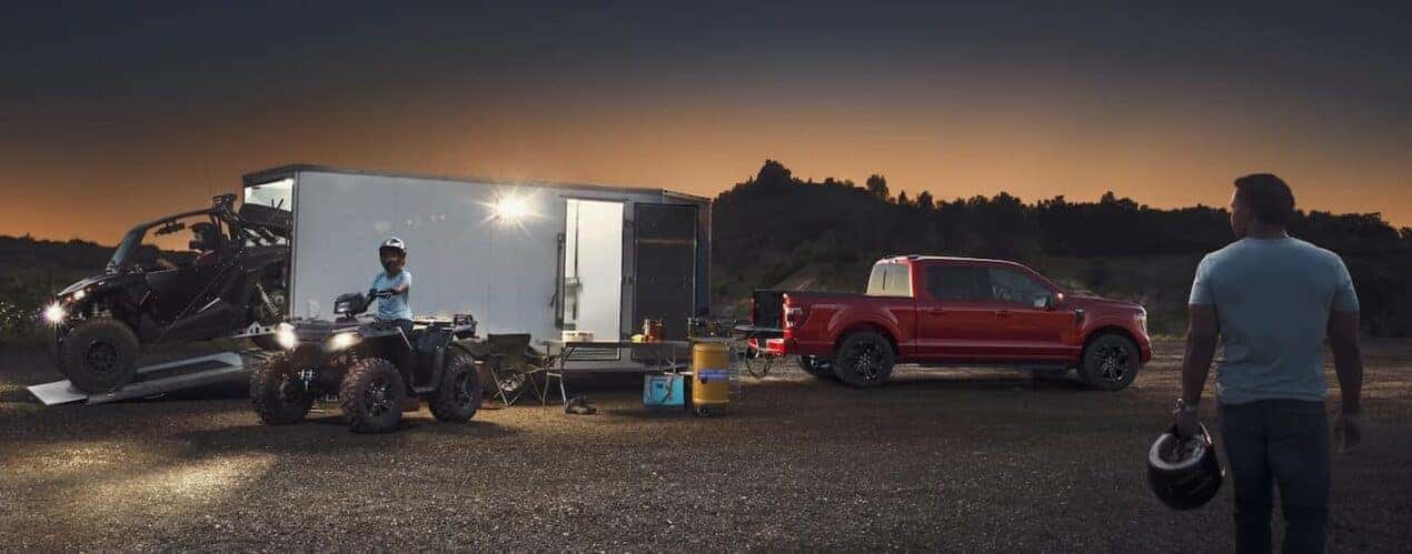 A red 2023 Ford F-150 is shown towing a trailer while surrounded by ATVs at sunset.