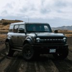 A black Ford Bronco driving on a muddy dirt road with mountains and cloudy skies in the background.