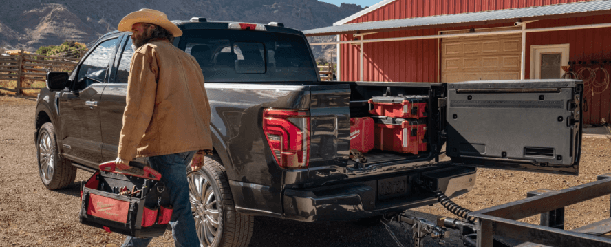 A man with a toolbox walking by a black Ford F-150 on a ranch with its truck bed open.