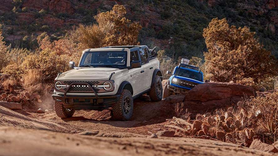 A silver and a blue Ford Bronco driving off-road through a bumpy desert.