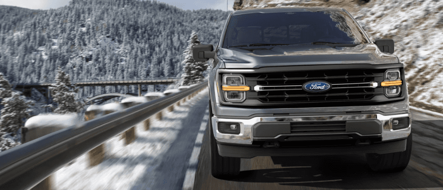 A front view of a gray Ford F-150 driving on the road around a snowy mountain.