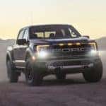 One of the most popular Ford performance vehicles, a black 2021 Ford Raptor, is shown parked on dirt with the headlights illuminated.